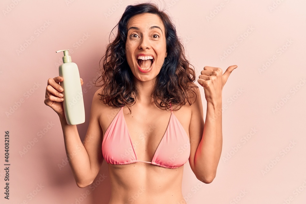 Young beautiful hispanic woman wearing bikini holding sunscreen lotion pointing thumb up to the side smiling happy with open mouth Young beautiful hispanic woman wearing bikini holding sunscreen lotion pointing thumb up to the side smiling happy with open mouth