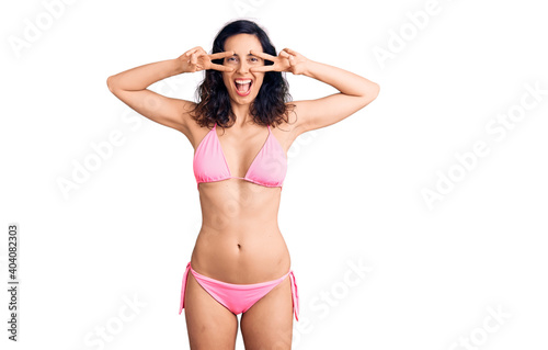 Young beautiful hispanic woman wearing bikini doing peace symbol with fingers over face, smiling cheerful showing victory © Krakenimages.com