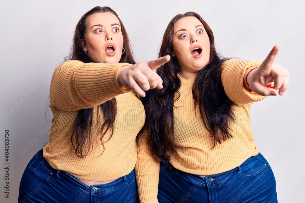 Young plus size twins wearing casual clothes pointing with finger surprised ahead, open mouth amazed expression, something on the front Young plus size twins wearing casual clothes pointing with finger surprised ahead, open mouth amazed expression, something on the front