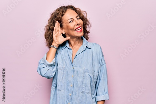 Canvastavla Middle age beautiful woman wearing casual denim shirt standing over pink background smiling with hand over ear listening an hearing to rumor or gossip