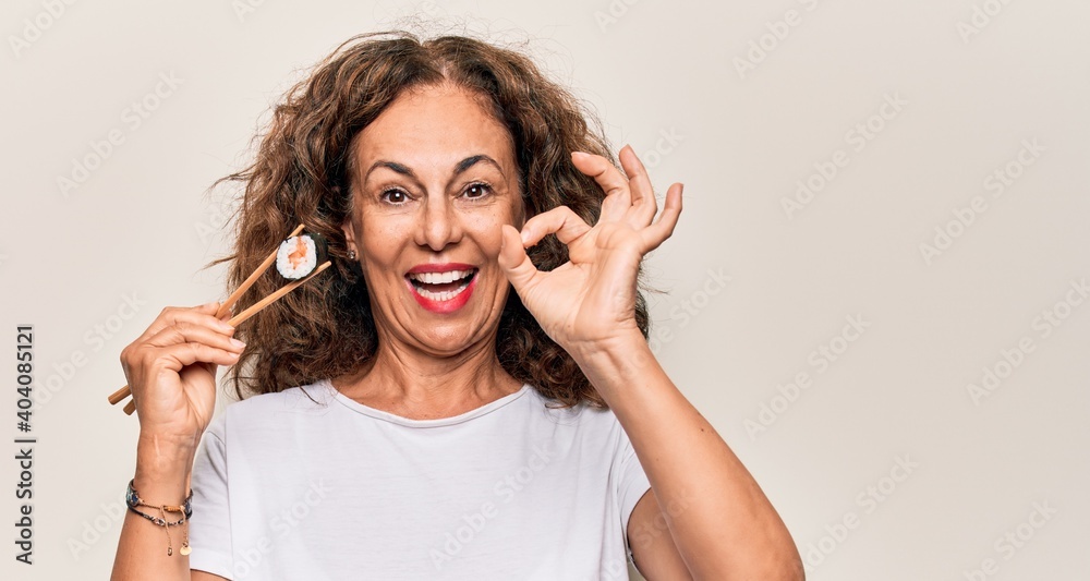 Middle age beautiful woman eating sushi using wooden chopsticks over white background doing ok sign with fingers, smiling friendly gesturing excellent symbol Middle age beautiful woman eating sushi using wooden chopsticks over white background doing ok sign with fingers, smiling friendly gesturing excellent symbol
