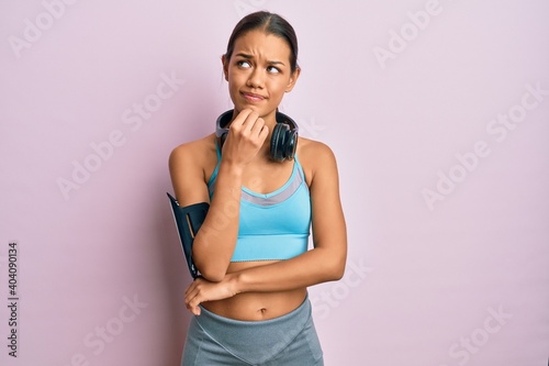 Beautiful hispanic woman wearing sportswear and headphones thinking concentrated about doubt with finger on chin and looking up wondering