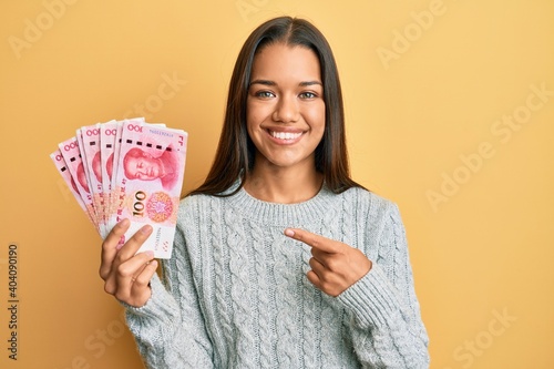 Beautiful hispanic woman holding 100 yuan chinese banknotes smiling happy pointing with hand and finger