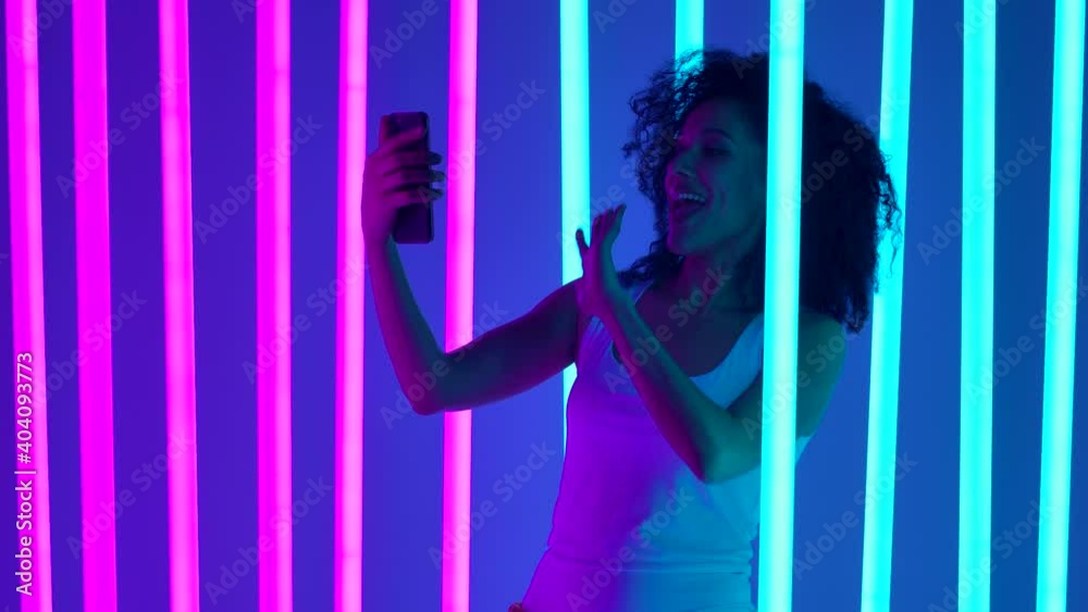 Portrait of a african american woman emotionally talking through a video call on her smartphone. The girl poses in the studio against the background of blue and pink neon lamps. Slow motion. Close up. Portrait of a african american woman emotionally talking through a video call on her smartphone. The girl poses in the studio against the background of blue and pink neon lamps. Slow motion. Close up.