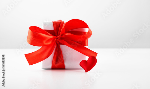 Gift box with red bow ribbon and red paper heart on white table for Valentines day.