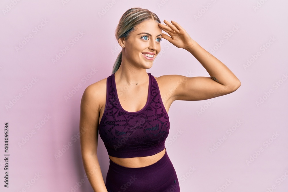Beautiful blonde woman wearing sportswear over pink background very happy and smiling looking far away with hand over head. searching concept. Beautiful blonde woman wearing sportswear over pink background very happy and smiling looking far away with hand over head. searching concept.