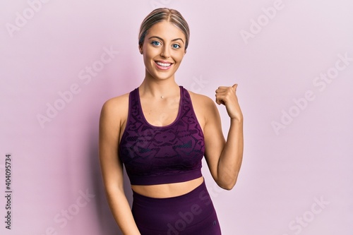 Beautiful blonde woman wearing sportswear over pink background smiling with happy face looking and pointing to the side with thumb up. © Krakenimages.com