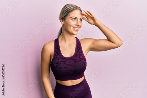 Beautiful blonde woman wearing sportswear over pink background very happy and smiling looking far away with hand over head. searching concept.