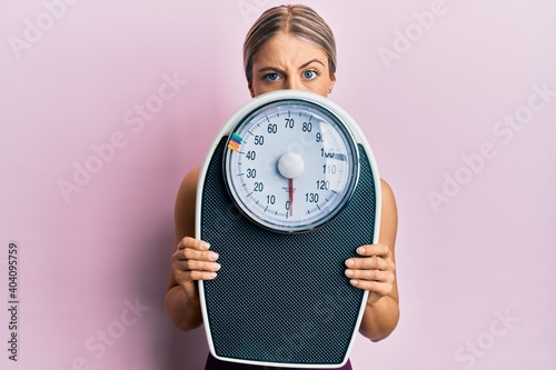 Konstfotografi Beautiful blonde woman holding weight machine to balance weight loss skeptic and nervous, frowning upset because of problem