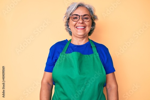 Senior hispanic woman wearing apron and glasses looking positive and happy standing and smiling with a confident smile showing teeth photo