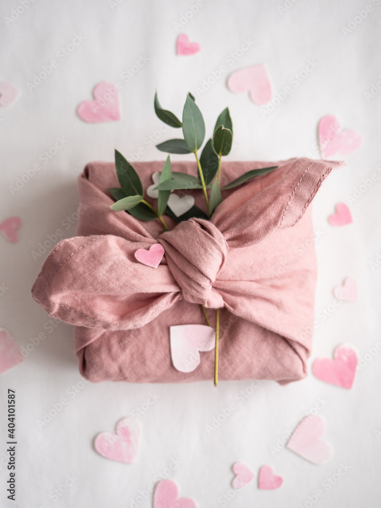 Gift wrapped in pink linen decorated with eucalyptus on a background of paper pink hearts on a white background in the morning light Gift wrapped in pink linen decorated with eucalyptus on a background of paper pink hearts on a white background in the morning light