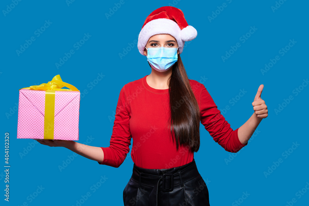 Obraz premium Young woman wearing Santa hat wearing face protective medical mask holding christmas or new year gift box and showing thumb up gesture, blue isolated background.