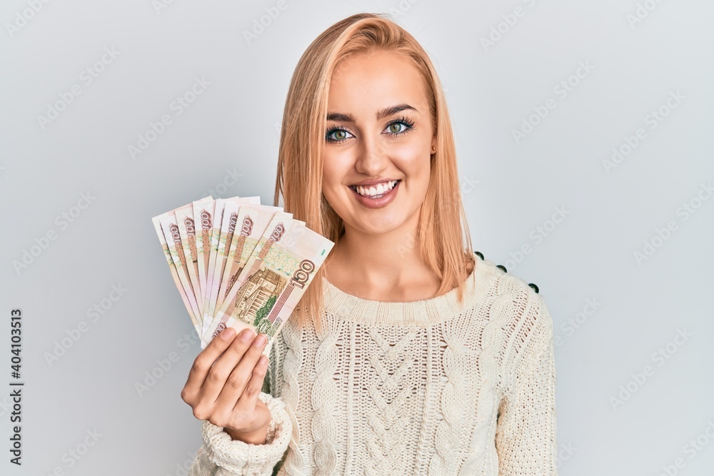 Beautiful caucasian woman holding 100 russian ruble banknotes looking positive and happy standing and smiling with a confident smile showing teeth Beautiful caucasian woman holding 100 russian ruble banknotes looking positive and happy standing and smiling with a confident smile showing teeth