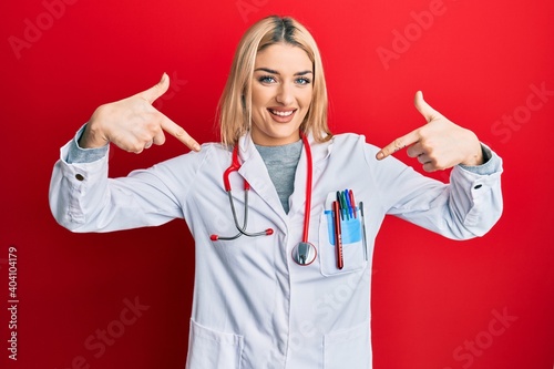 Young caucasian woman wearing doctor uniform and stethoscope looking confident with smile on face  pointing oneself with fingers proud and happy.
