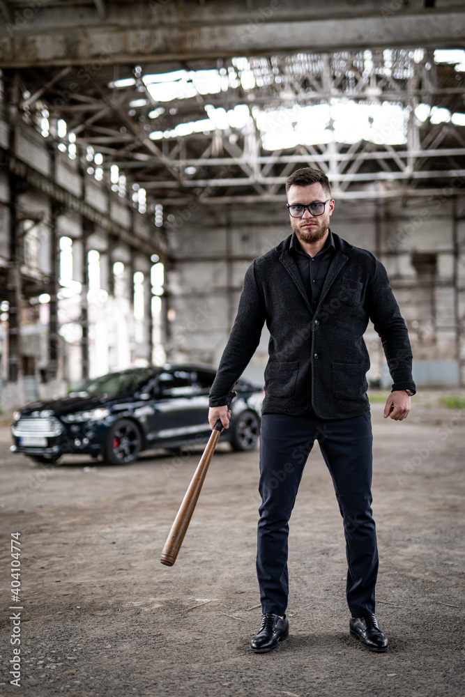 Full lenght portrait of a cool brutal guy with baseball bat in hands and modern car on the background. Total black outfit with sunglasses.