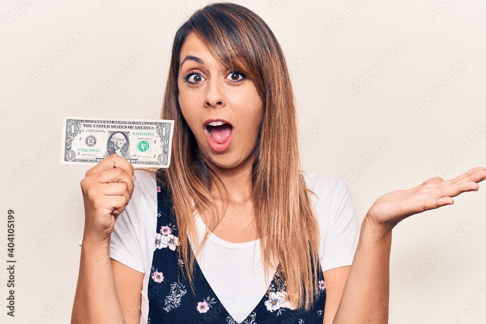 Young beautiful woman holding one dollar celebrating achievement with happy smile and winner expression with raised hand Young beautiful woman holding one dollar celebrating achievement with happy smile and winner expression with raised hand