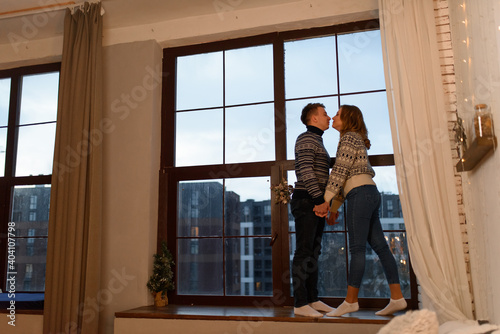 A loving couple in knitted sweaters stand by the hand against the background of a large window on the windowsill.