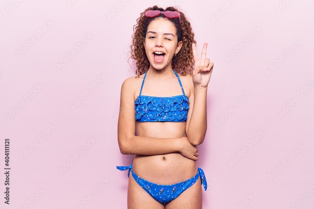 Naklejka premium Beautiful kid girl with curly hair wearing bikini and sunglasses smiling with happy face winking at the camera doing victory sign. number two.