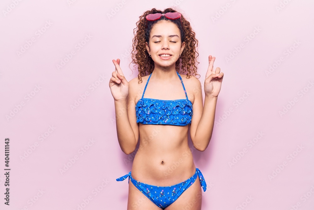 Naklejka premium Beautiful kid girl with curly hair wearing bikini and sunglasses gesturing finger crossed smiling with hope and eyes closed. luck and superstitious concept.