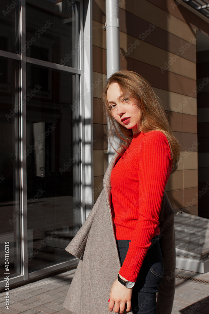Portrait of a young woman taking off coat, looking at camera and standing on street in front of window and brown wall. Caucasian girl having fun outside. Urban style and fashion. Casual outfit Portrait of a young woman taking off coat, looking at camera and standing on street in front of window and brown wall. Caucasian girl having fun outside. Urban style and fashion. Casual outfit