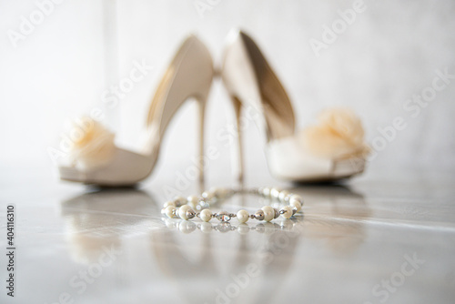 Pearl bracet in front of High heel bridal shoes with silk flowers on reflective surface