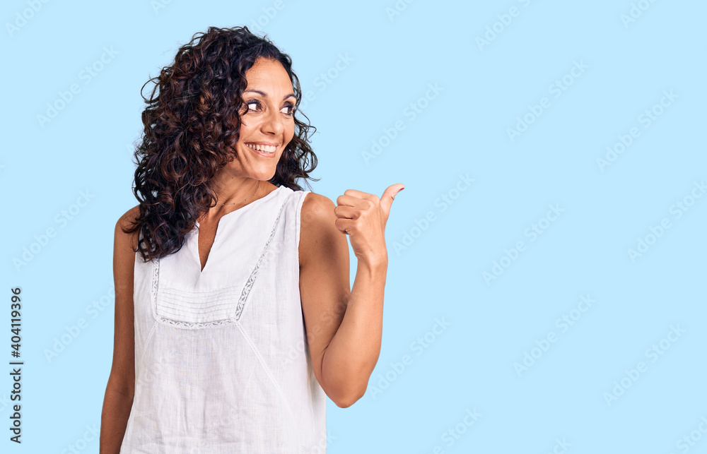 Middle age beautiful woman wearing casual sleeveless t shirt smiling with happy face looking and pointing to the side with thumb up. Middle age beautiful woman wearing casual sleeveless t shirt smiling with happy face looking and pointing to the side with thumb up.