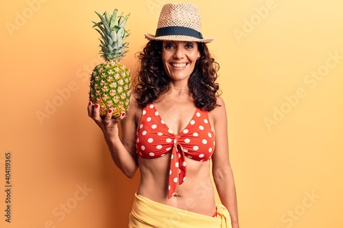 Middle age beautiful woman wearing bikini and summer hat holding pineappel looking positive and happy standing and smiling with a confident smile showing teeth photo