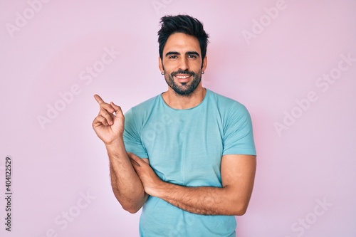 Handsome hispanic man with beard wearing casual clothes with a big smile on face, pointing with hand and finger to the side looking at the camera.