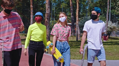 Stylish group of friends guys and ladies in the pandemic of Coronavirus in the modern skate park wearing protective mask they walking and holding their skateboard one of the guy skateboard beside photo