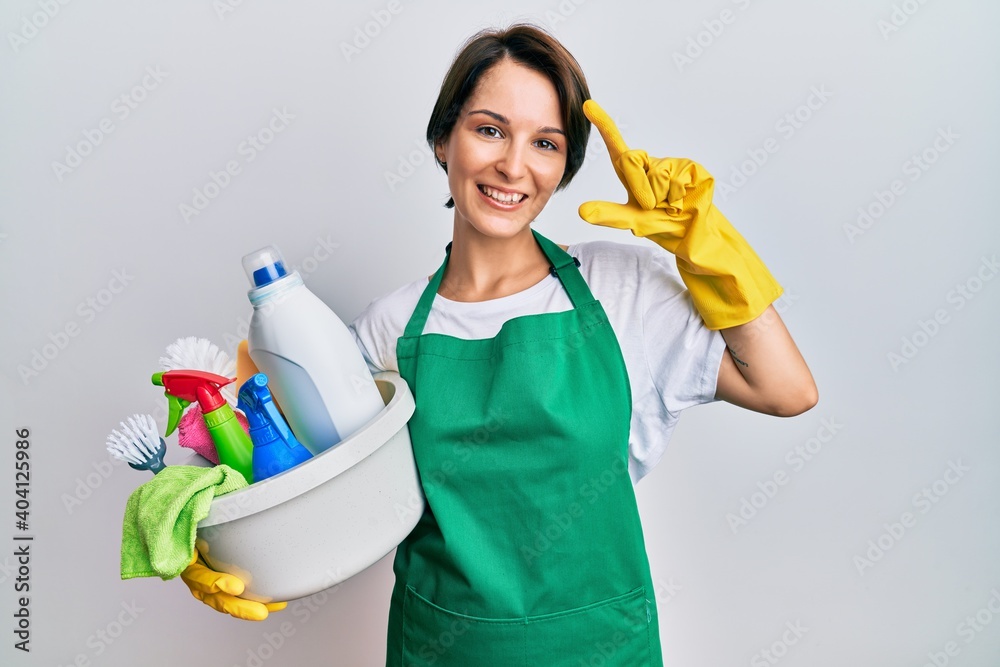Young brunette woman with short hair wearing apron holding cleaning products smiling and confident gesturing with hand doing small size sign with fingers looking and the camera. measure concept. Young brunette woman with short hair wearing apron holding cleaning products smiling and confident gesturing with hand doing small size sign with fingers looking and the camera. measure concept.