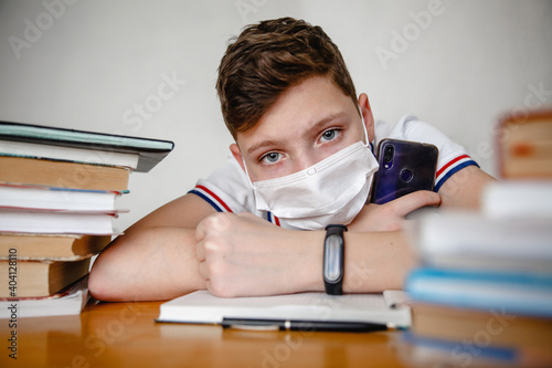 Portrait of a teenager at home in a mask behind textbooks. In the hands of a smartphone. Coronavirus concept