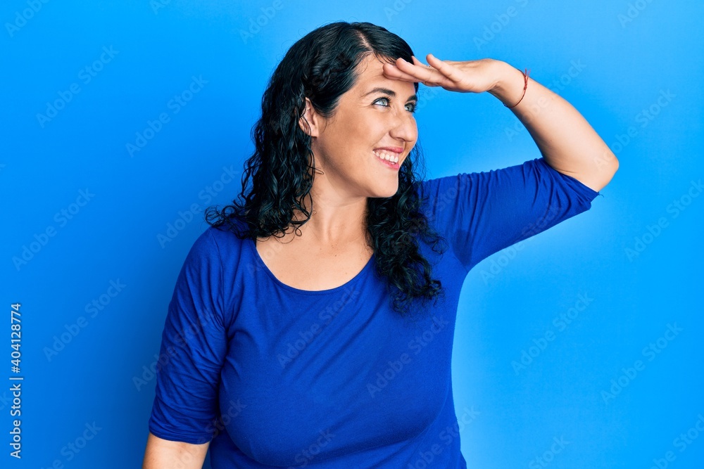 Plus size brunette woman wearing casual blue shirt very happy and smiling looking far away with hand over head. searching concept. Plus size brunette woman wearing casual blue shirt very happy and smiling looking far away with hand over head. searching concept.