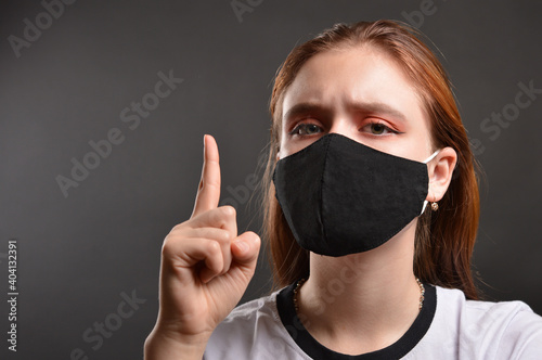 Close-up of a young girl in a medical mask with a raised index finger against a dark background.