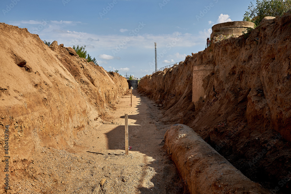 A wooden stake for setting the height of the base of the pipeline laying in  the trench. A trench for installing water pipes leading to the sleeve  across the road Photos