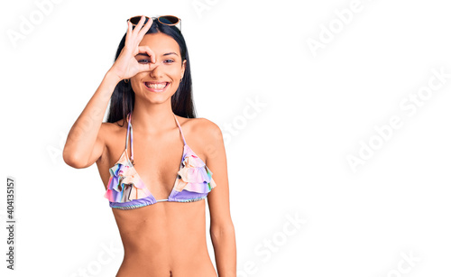 Young beautiful latin girl wearing bikini and sunglasses doing ok gesture with hand smiling, eye looking through fingers with happy face. © Krakenimages.com