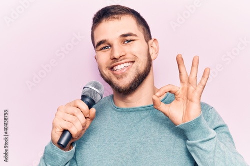 Young handsome man singing song using microphone doing ok sign with fingers, smiling friendly gesturing excellent symbol