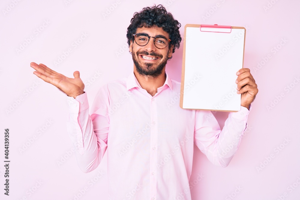 Handsome young man with curly hair and bear holding clipboard with blank space celebrating victory with happy smile and winner expression with raised hands Handsome young man with curly hair and bear holding clipboard with blank space celebrating victory with happy smile and winner expression with raised hands