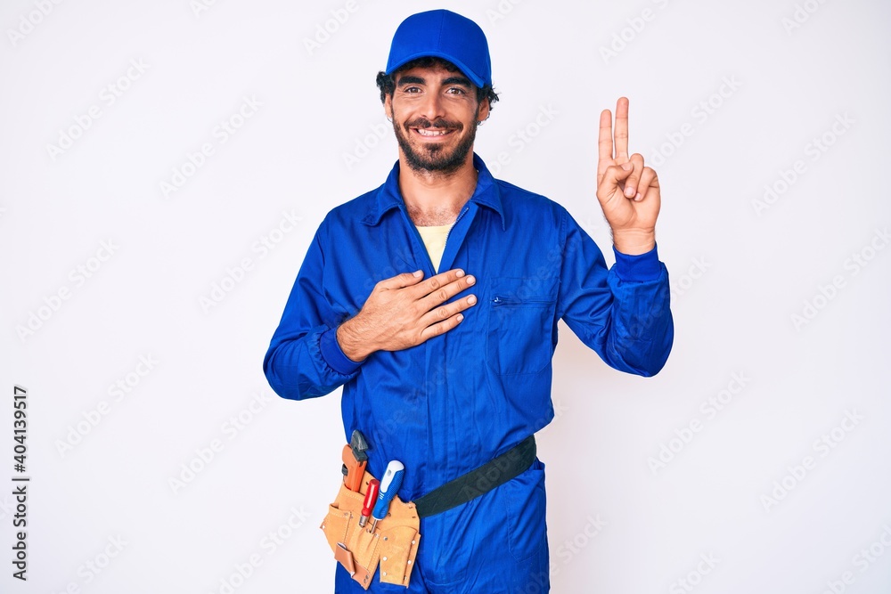 Handsome young man with curly hair and bear weaing handyman uniform smiling swearing with hand on chest and fingers up, making a loyalty promise oath