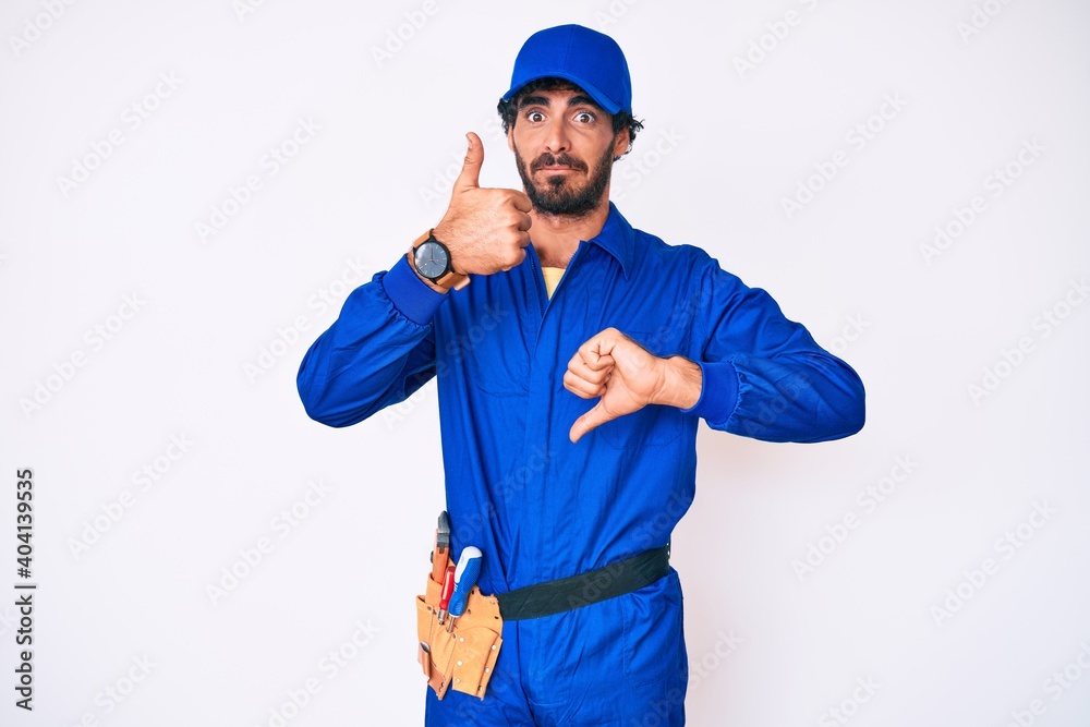 Handsome young man with curly hair and bear weaing handyman uniform doing thumbs up and down, disagreement and agreement expression. crazy conflict