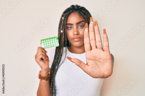 Young african american woman with braids holding birth control pills with open hand doing stop sign with serious and confident expression, defense gesture