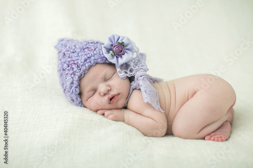 Serene sleeping newborn baby girl with a purple lavender crochet elf type pointed hat on her head curled up on a cream-colored blanket