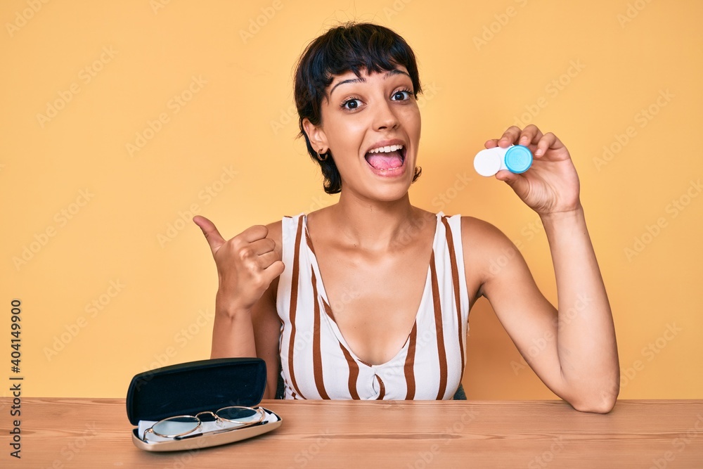 Beautiful brunettte woman holding contact lenses pointing thumb up to the side smiling happy with open mouth Beautiful brunettte woman holding contact lenses pointing thumb up to the side smiling happy with open mouth