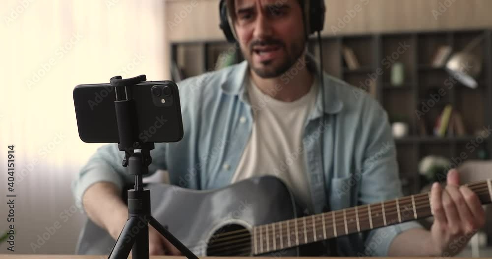 Young male blogger wear headphones playing acoustic guitar string instrument recording music video sing own song, vlogger artist create tutorial on smartphone at home studio. Hobby, streaming concept Young male blogger wear headphones playing acoustic guitar string instrument recording music video sing own song, vlogger artist create tutorial on smartphone at home studio. Hobby, streaming concept