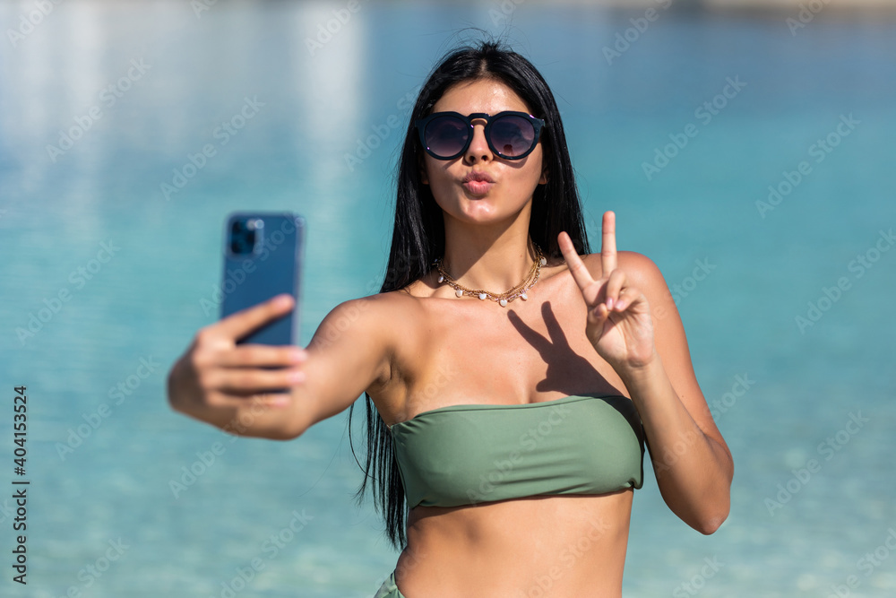 Beautiful young woman doing selfie with peace gesture on the beach