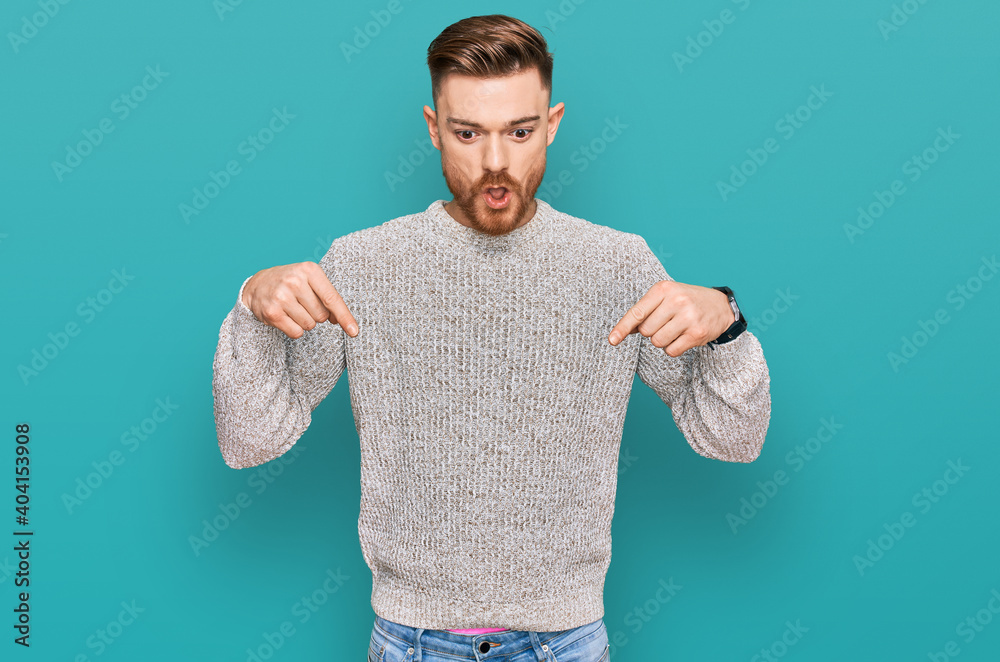 Young redhead man wearing casual winter sweater pointing down with fingers showing advertisement, surprised face and open mouth Young redhead man wearing casual winter sweater pointing down with fingers showing advertisement, surprised face and open mouth