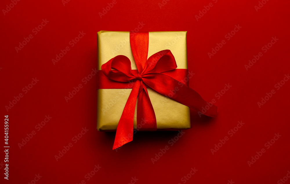 Holiday gift. Box in a gold gift wrapping with a red bow, on a red background. Merry Christmas, Happy New Year and Valentine's Day greetings. Postcard to loved ones. High quality photo