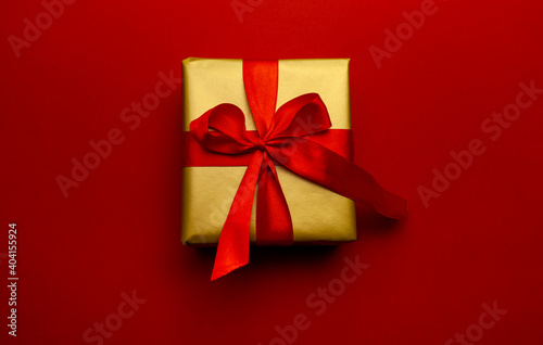 Holiday gift. Box in a gold gift wrapping with a red bow, on a red background. Merry Christmas, Happy New Year and Valentine's Day greetings. Postcard to loved ones. High quality photo