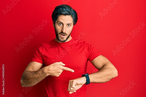 Young hispanic man wearing casual red t shirt in hurry pointing to watch time, impatience, upset and angry for deadline delay