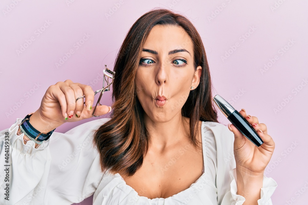 Young brunette woman holding eyelash curler making fish face with mouth and squinting eyes, crazy and comical. Young brunette woman holding eyelash curler making fish face with mouth and squinting eyes, crazy and comical.