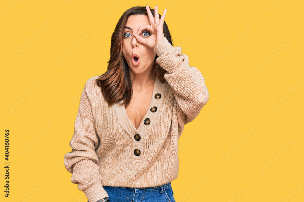 Young brunette woman wearing casual winter sweater doing ok gesture shocked with surprised face, eye looking through fingers. unbelieving expression.